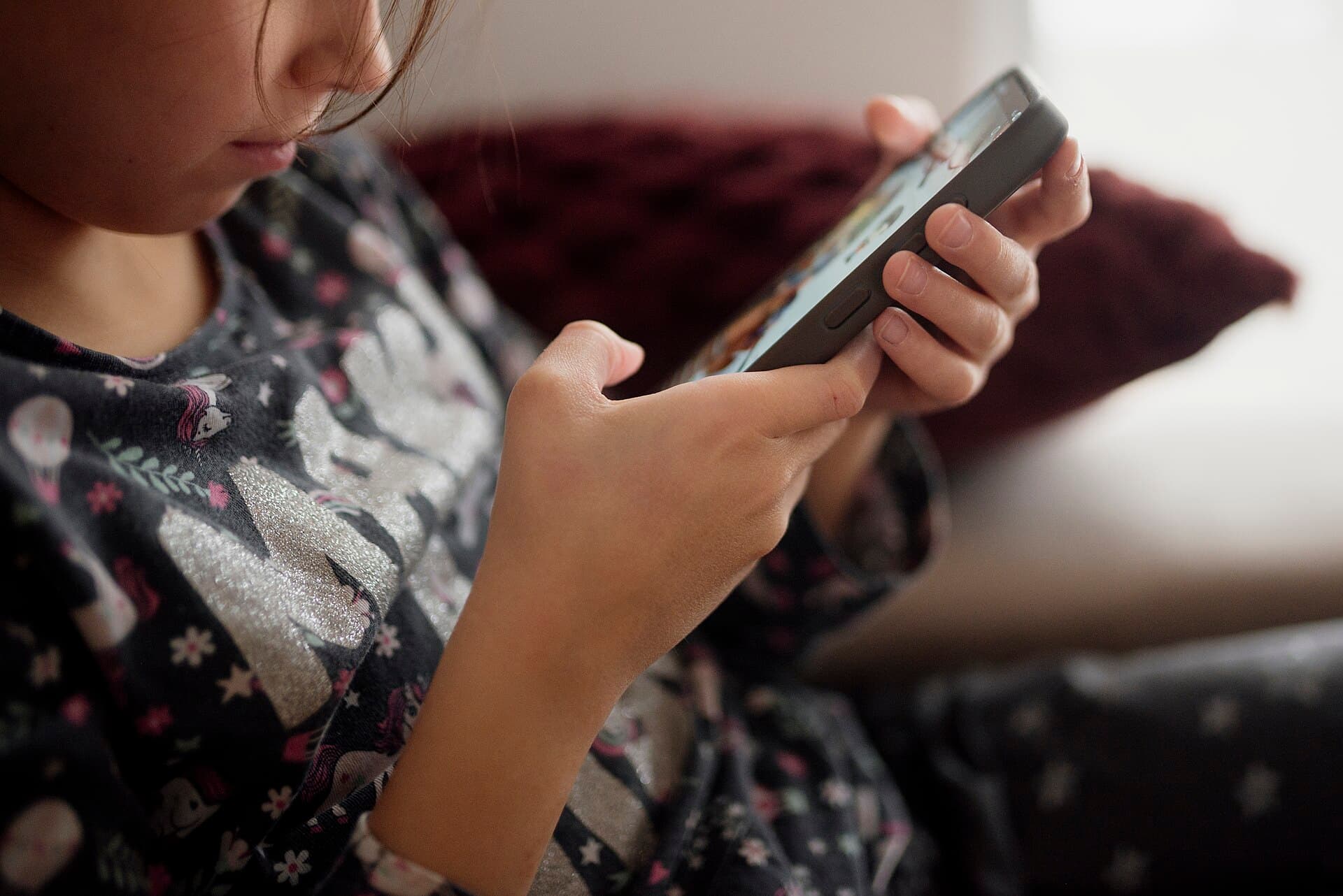 Child plays on smartphone while sitting on a couch during afternoon at home in cozy living room setting