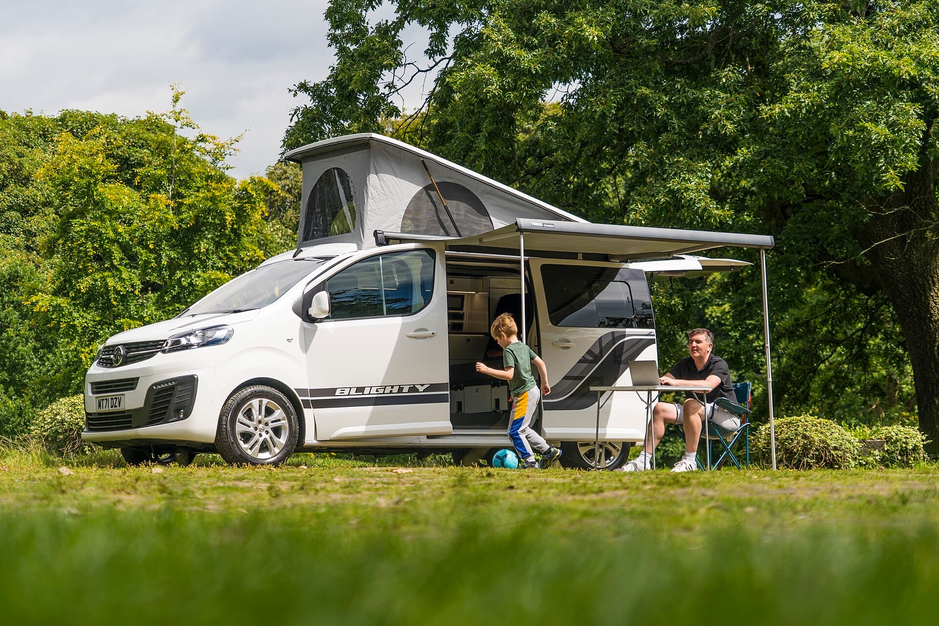 A child plays soccer beside a camper van while his father works remotely