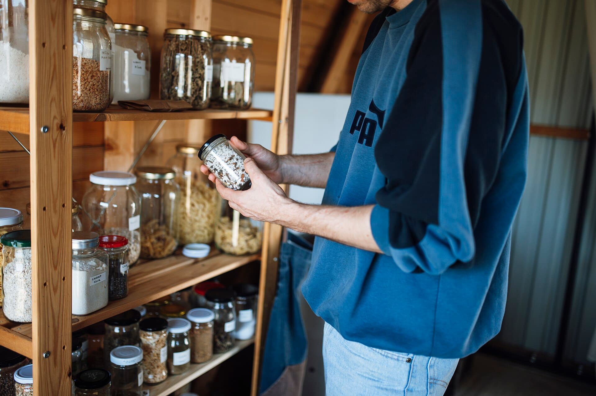 Person examines a jar of spice on a wooden shelf in a kitchen pantry during daytime in a cozy home setting