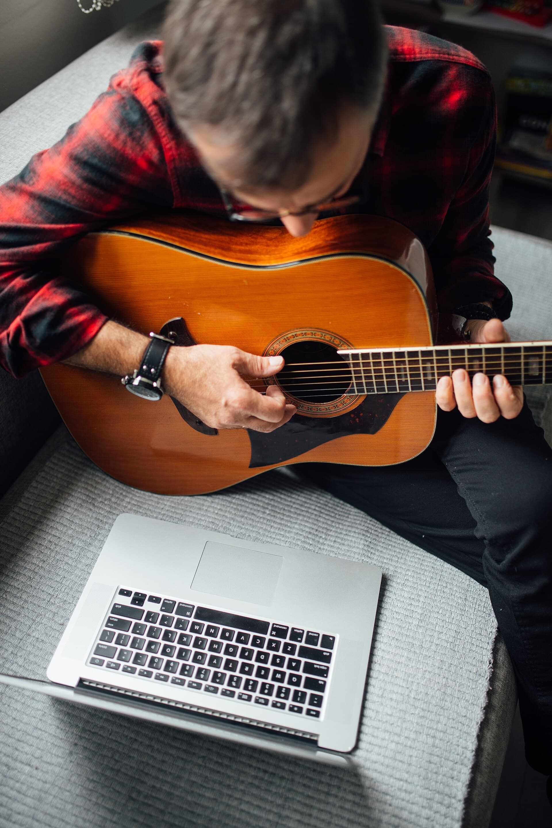 Person playing guitar at home while using laptop for music inspiration in a cozy indoor setting